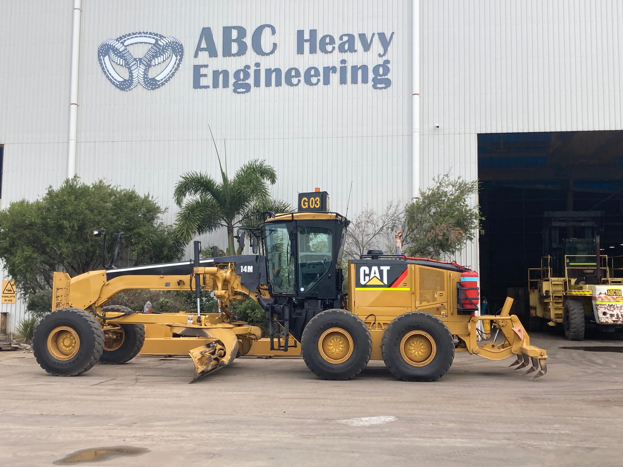 Caterpillar 14M grader parked in front of the signed ABC Heavy Engineering workshop, heavy forklift ABC108 in the doorway
